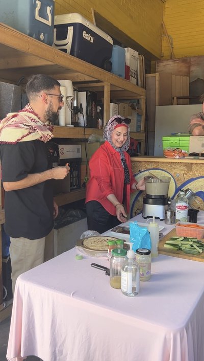 two people with a blender, fresh ingredients, smiling and making food. The man is wearing a keffiyeh, and the woman has a colourful shirt and hijab, excitedly cooking