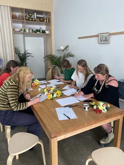five people sitting at a table writing letters. There are colourful flowers next to them