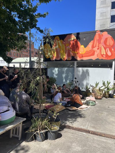 succulents, trees and people sitting on the ground in the shade. There's a olourful mural on a sunny day
